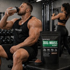 Muscular man sitting in a gym drinking a protein shake, with a HEROES XXL-MASS PLANT chocolate protein powder bag displayed prominently beside him and a woman working out in the background